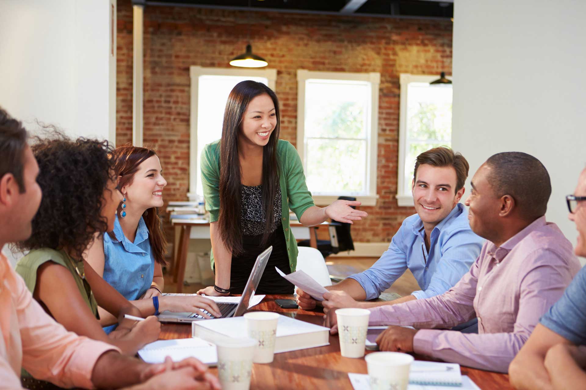 A diverse group of people are happily discussing in a brightly lit room. Some are holding papers and one has a laptop.